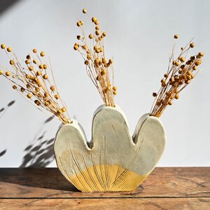 May include: A three-opening ceramic vase with a light gray upper section and a yellow base featuring etched vertical lines. The vase contains three bundles of dried brown flax stems with small round seed pods extending from the openings.