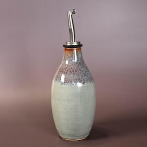 May include: A ceramic olive oil bottle with a stainless steel spout. The bottle has a light green base, transitioning to a brown and gray speckled top. The spout is curved and has a small opening.