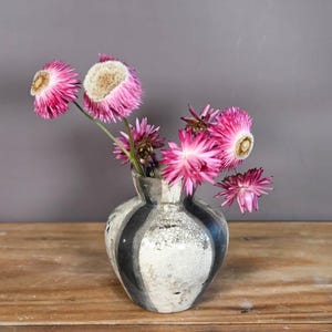 May include: A vase with a black and white striped design displays a collection of pink and white dried flowers. The vase is placed on a wooden surface, set against a grey backdrop.