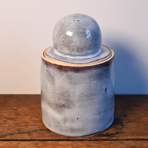 Puede incluir: Un tarro de cerámica con tapa. El tarro es cilíndrico con un esmalte moteado gris y blanco. La tapa es una esfera redondeada con un esmalte moteado a juego. El borde del tarro tiene una banda roja oscura. El tarro está sobre una superficie de madera.