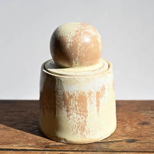 May include: A handmade ceramic jar with a lid. The jar is cylindrical with a rounded top and a spherical lid. The colors are a mix of beige and off-white, with a textured, dripped glaze effect. The jar sits on a wooden surface.
