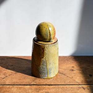 May include: A cylindrical ceramic jar with a spherical lid, featuring a mottled glaze in shades of mustard yellow, olive green, and earthy brown. The surface has a textured, uneven finish with visible drips and variations in the glaze color.
