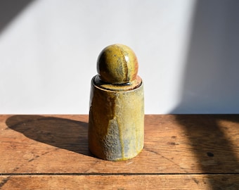 May include: A cylindrical ceramic jar with a spherical lid, featuring a mottled glaze in shades of mustard yellow, olive green, and earthy brown. The surface has a textured, uneven finish with visible drips and variations in the glaze color.
