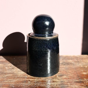 Könnte beinhalten: Ein dunkelblaues Keramikgefäß mit rundem Deckel. Das Gefäß hat einen zylindrischen Körper und steht auf einer Holzoberfläche. Die Oberfläche des Gefäßes hat ein gesprenkeltes Aussehen, und der Deckel ist dunkler blau. Das Gefäß ist etwa 15 cm hoch.