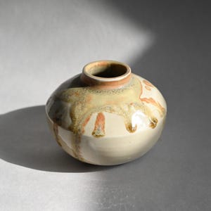 May include: A small, round ceramic vase with a white glaze and brown and orange drips. The vase has a wide opening and a rounded bottom.