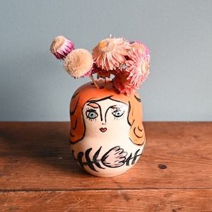 May include: A ceramic vase shaped like a woman's face with orange hair, black eyes, and a pink flower on her chest. The vase is decorated with black lines and is holding dried pink flowers.