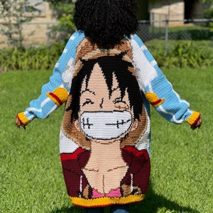 May include: A handmade, crochet cardigan featuring a large anime character design on the back. The cardigan has a blue, white, red, yellow, and brown color scheme. The sleeves have a blue and white cloud pattern with yellow and red accents.