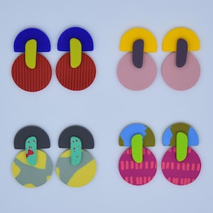 Puede incluir: Una colección de pendientes abstractos y coloridos. Cada par presenta una combinación única de formas y colores, incluyendo rojo, azul, amarillo, rosa y verde. Los pendientes tienen un diseño moderno y geométrico, con elementos sólidos y estampados.