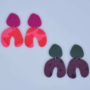 May include: Two pairs of abstract earrings. One pair has a fuchsia top and a red and pink patterned bottom. The other pair has a dark green top and a dark purple bottom with a speckled design. The earrings are connected by a small purple loop.