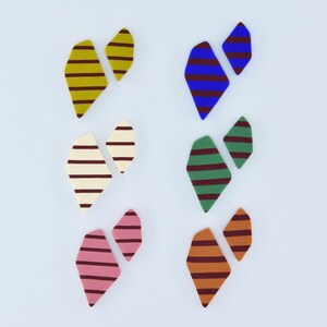Può includere: Sei paia di oggetti astratti a forma di cuore in vari colori, ciascuno con strisce orizzontali. I colori includono giallo, blu, verde, crema, rosa e arancione, tutti con strisce marroni. Gli oggetti sono disposti su uno sfondo bianco.