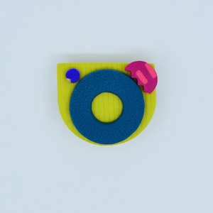 Op de afbeelding: Abstracte broche met een gele, halfronde basis, een blauwe cirkel en een roze, halfronde vorm. Er is ook een kleine blauwe cirkel aanwezig. De broche staat tegen een witte achtergrond.