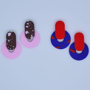 Pode incluir: Dois pares de brincos. Um com um círculo rosa e um oval marrom salpicado de branco. O outro com um oval vermelho e um círculo azul com manchas vermelhas. Fundo azul claro.