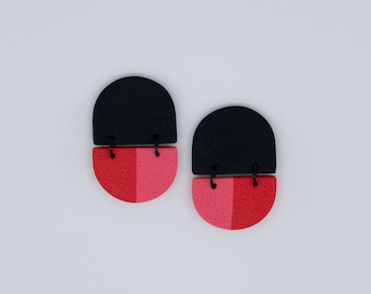 Peut inclure: Une paire de boucles d'oreilles géométriques noires et roses. Les boucles d'oreilles sont composées de deux pièces, un demi-cercle noir et un demi-cercle rose. Les deux pièces sont reliées par une ficelle noire.