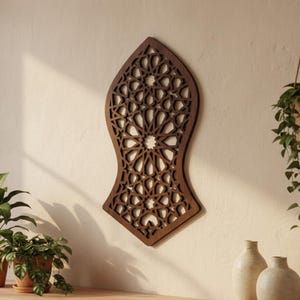 Peut inclure: Une œuvre d'art murale en bois brun foncé, finement sculptée avec un motif géométrique, fixée sur un mur beige. Le design présente un motif central en étoile et une série de formes répétitives. L'œuvre est exposée au-dessus d'un meuble en bois avec des plantes en pot et des vases décoratifs.