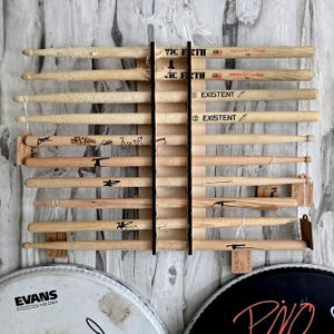 Pode incluir: Um expositor de baquetas de madeira com várias baquetas. As baquetas são de cor clara e estão dispostas horizontalmente. Algumas têm texto, incluindo "VIC FIRTH" e "EXISTENT". O expositor está montado numa parede texturizada de cor clara.