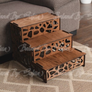 以下が含まれることがあります： 茶色のステイン仕上げの3段木製ペット用階段。各段には、抽象的な形のカットアウトデザインが施されています。階段は茶色のソファの前に置かれています。