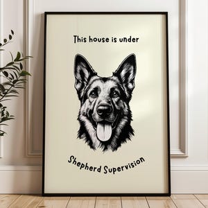May include: Black framed print featuring a detailed black and white illustration of a German Shepherd dog. The text "This house is under Shepherd Supervision" is above and below the dog's image. The artwork is displayed on a light beige background.