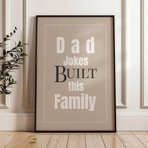Puede incluir: Arte mural enmarcado con el texto "Dad Jokes Built This Family" en fuentes blancas y gris oscuro. La obra de arte está sobre un fondo beige, enmarcada en negro y expuesta sobre un suelo de madera.