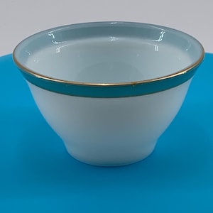 May include: A white ceramic bowl with a light blue rim and a thin gold band around the edge. The bowl is sitting on a bright blue surface.