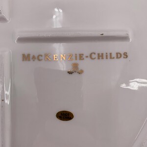 May include: A white ceramic surface featuring the gold text MACKENZIE-CHILDS with a small gold thistle emblem centered below. An oval gold sticker with the black text MADE IN CHINA is positioned near the bottom of the white ceramic piece.