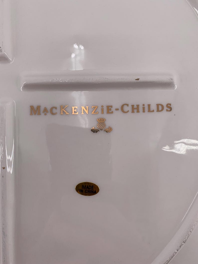 May include: A white ceramic surface featuring the gold text MACKENZIE-CHILDS with a small gold thistle emblem centered below. An oval gold sticker with the black text MADE IN CHINA is positioned near the bottom of the white ceramic piece.