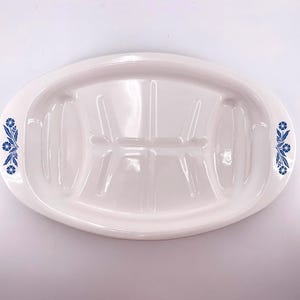 May include: An oval white ceramic serving platter featuring a central grooved drainage pattern and blue floral motifs on both side handles. The platter has a smooth, glossy finish and a wide rim, designed for presenting food with integrated channels.