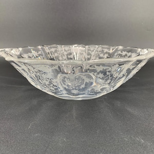 May include: Clear glass bowl with a scalloped rim and etched floral and bird designs. The bowl has a wide, shallow shape and is likely used for serving or display. The glass is transparent, allowing light to pass through.