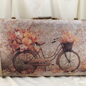 May include: Vintage suitcase with a floral bicycle design. The suitcase features a brown bicycle with a basket of flowers and a background of flowers in shades of peach, orange, and yellow. The suitcase has a cream-colored frame and gold-colored hardware.