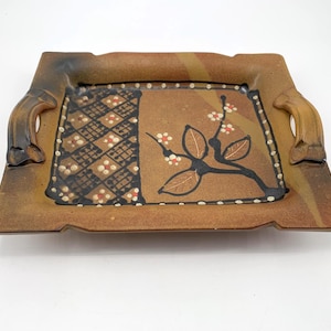 May include: A square, ceramic serving tray with integrated handles. The tray features a brown and tan color scheme with a decorative floral design in the center. The design includes a black branch with leaves and small white and red flowers.