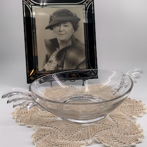 May include: A clear glass bowl with two handles in the shape of stylized leaves. The bowl is sitting on a crocheted doily. A black framed photo of a woman wearing a hat is in the background.