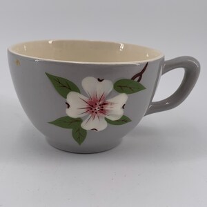 May include: A gray ceramic teacup with a white flower and green leaves painted on the side. The cup has a rounded shape and a handle.