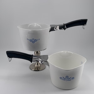 May include: Two white ceramic saucepans with blue floral designs and black handles. The saucepans have a silver metal base and a silver metal clip that attaches the handle to the saucepan.