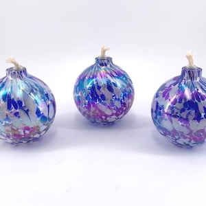May include: Three decorative glass oil lamps with a speckled design in shades of blue, purple, and silver. Each lamp has a wick and a round, bulbous shape, perfect for adding ambiance to any space.
