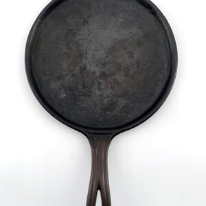 May include: A black cast iron griddle pan with a round cooking surface and a long handle with a hole for hanging. The pan is dark with signs of use, suggesting it is well-seasoned. The handle is a dark brown color.