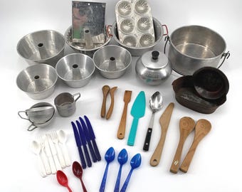 May include: A collection of vintage kitchen utensils and cookware, including aluminum bundt pans, a muffin tin, a small teapot, wooden spoons, and various serving spoons and knives. The utensils are in various colors, including blue, red, and white.