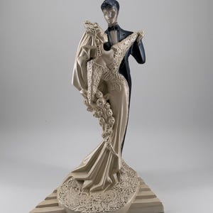 May include: A decorative figurine of a couple dancing. The woman wears a long, cream-colored gown with lace detailing and a veil. The man is in a black tuxedo. The base is a light beige with a geometric design.