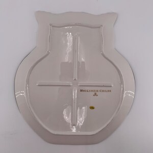 May include: The underside of an owl-shaped ceramic plate in a glossy off-white finish. The back features a raised cross-shaped support structure, the gold-colored text MACKENZIE-CHILDS, a small gold logo, and a tiny yellow circular sticker.