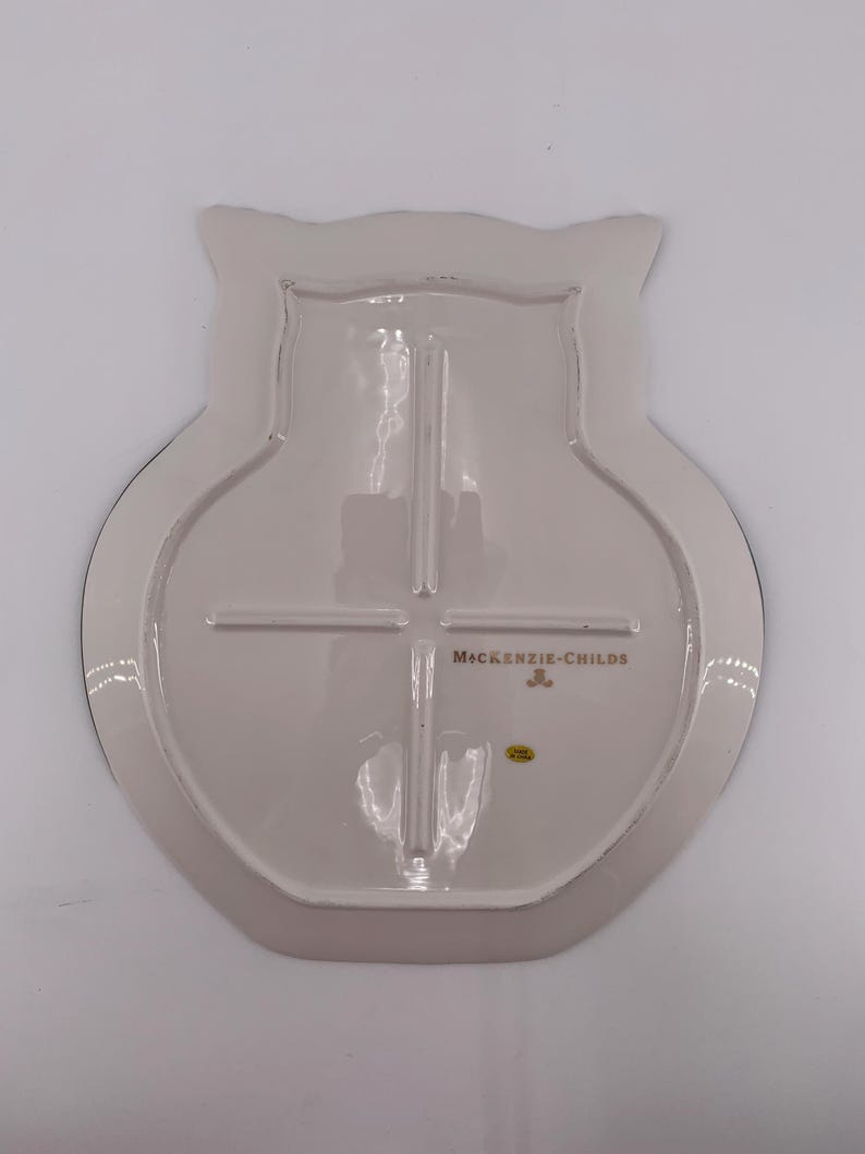 May include: The underside of an owl-shaped ceramic plate in a glossy off-white finish. The back features a raised cross-shaped support structure, the gold-colored text MACKENZIE-CHILDS, a small gold logo, and a tiny yellow circular sticker.