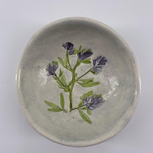 May include: A small, white ceramic bowl with a pressed flower design. The flower is purple and green and has a delicate, natural look.