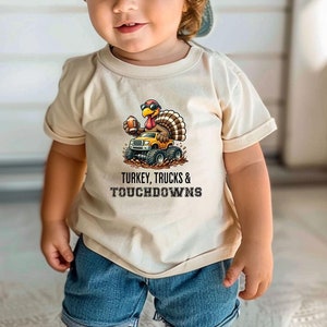 May include: A young child wearing a beige t-shirt and blue denim shorts. The t-shirt has a graphic of a turkey driving a monster truck with the text "TURKEY, TRUCKS & TOUCHDOWNS". The child is also wearing a light blue baseball cap.