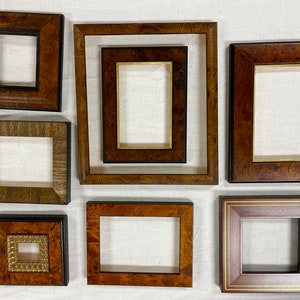 May include: An assortment of seven wooden picture frames in various sizes and styles. The frames feature rich brown tones, some with black or gold accents. The collection includes both square and rectangular shapes, showcasing different wood grain patterns.