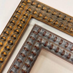 Peut inclure: Gros plan sur deux cadres photo décoratifs. L'un est doré avec des accents noirs, l'autre est gris avec des accents marron. Les deux cadres ont un motif texturé répétitif de formes arrondies sur les bords.