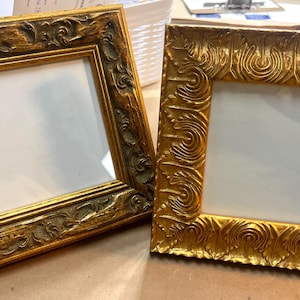 May include: Two ornate, square picture frames with gold-colored, detailed carvings. The frames have a distressed finish and white interiors. The frames are empty, ready to display photos or artwork.