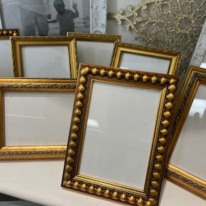 May include: A set of eight gold picture frames with ornate detailing. The frames are arranged in a staggered pattern on a white surface.