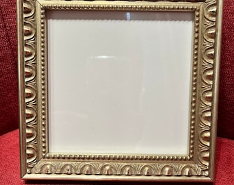 Puede incluir: Un marco de fotos cuadrado y ornamentado con acabado dorado. El marco tiene un diseño detallado y texturizado con un borde interior con cuentas y un borde superior festoneado. El marco está vacío, con un interior blanco.