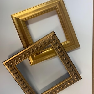 Puede incluir: Dos marcos de fotos dorados, uno con un diseño simple y el otro con un diseño más ornamentado, se colocan sobre una superficie blanca.