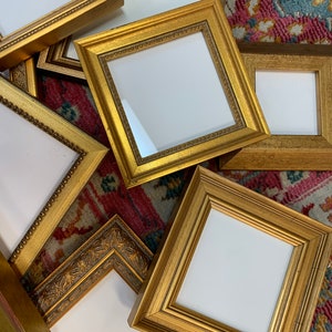 Puede incluir: Una colección de seis marcos de fotos dorados de varios tamaños y estilos, dispuestos sobre una alfombra estampada. Los marcos son todos de madera y tienen un acabado dorado. Algunos de los marcos tienen detalles ornamentados, mientras que otros son más simples.