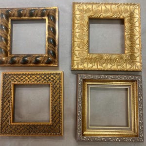 Op de afbeelding: Vier sierlijke fotolijsten in verschillende goud- en bronskleuren. De lijsten hebben gedetailleerde ontwerpen, waaronder touw, krullen en geometrische patronen. Elke lijst heeft een vierkante opening, klaar voor kunstwerken of foto's.