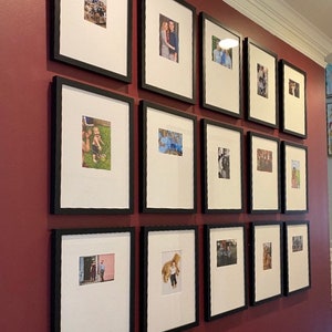 May include: A wall gallery of framed photographs in black frames on a burgundy wall. The photos are of various sizes and feature different subjects, including people, landscapes, and pets.