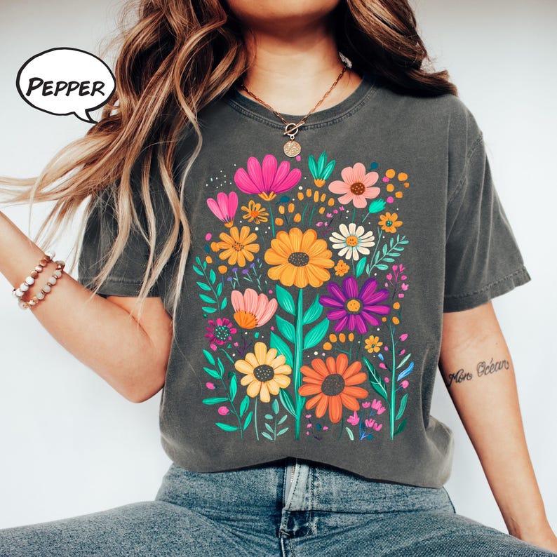 K&ouml;nnte beinhalten: Dunkelgraues T-Shirt mit einem lebendigen Blumendesign mit rosa, orangefarbenen, gelben und lila Blumen. Das Wort "PEPPER" befindet sich in einer Sprechblase. Das Shirt hat kurze &Auml;rmel und eine lockere Passform.